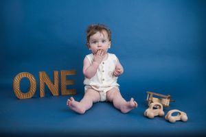 Baby celebrating first birthday milestone with wooden number ONE and teddy bear against blue backdrop.