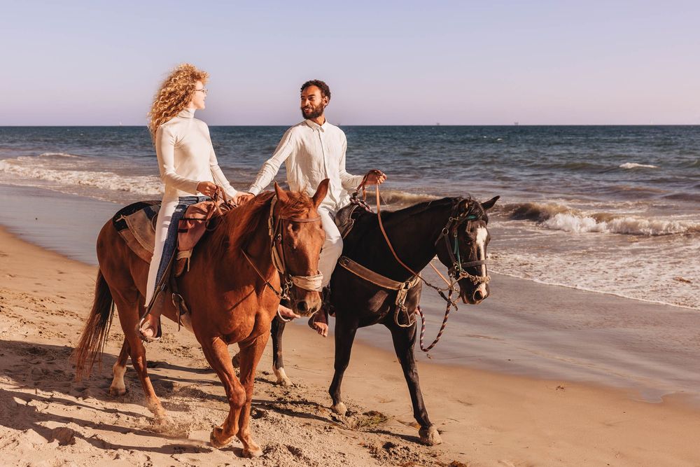 Two people horseback riding along a sandy beach at sunset with waves crashing on the shore.