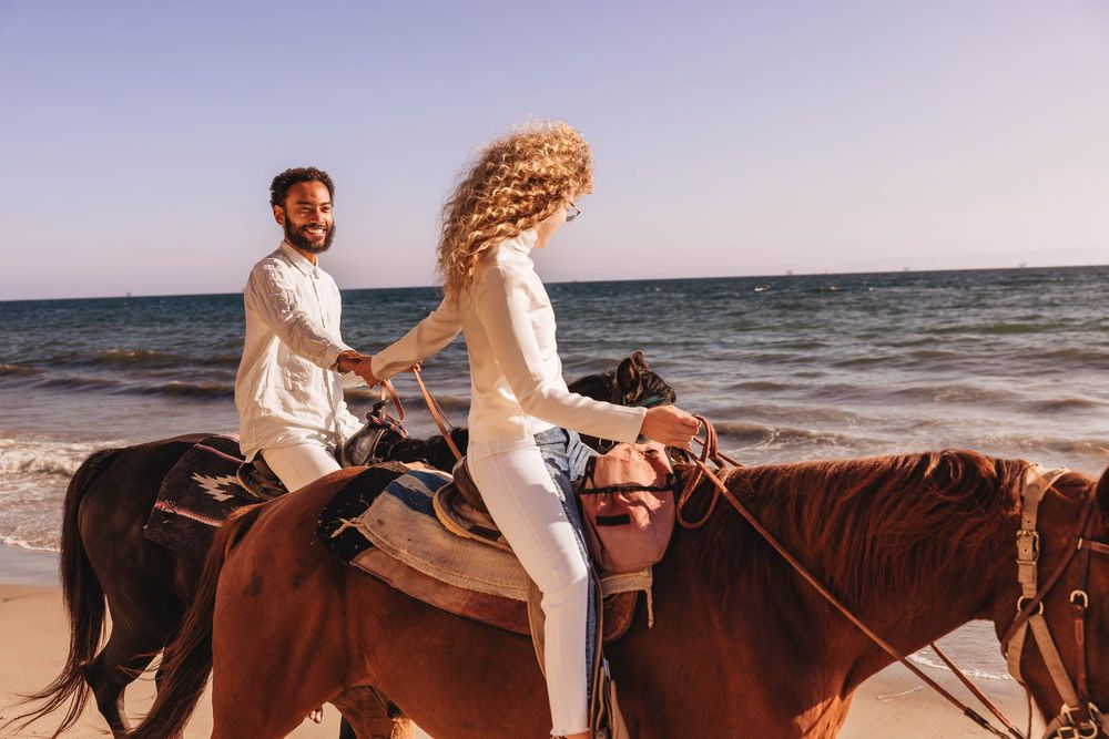 Couple riding horses together along a scenic beach during sunset, creating a romantic silhouette against the ocean.