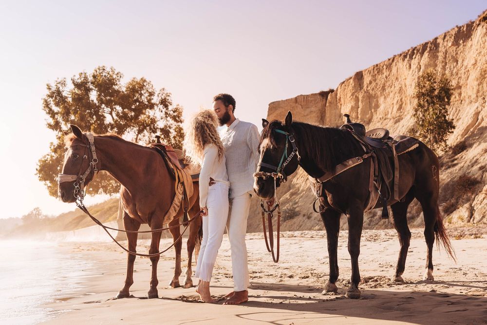 Two horses stand with couple in white outfits on a beach at sunset with cliffs in the background.
