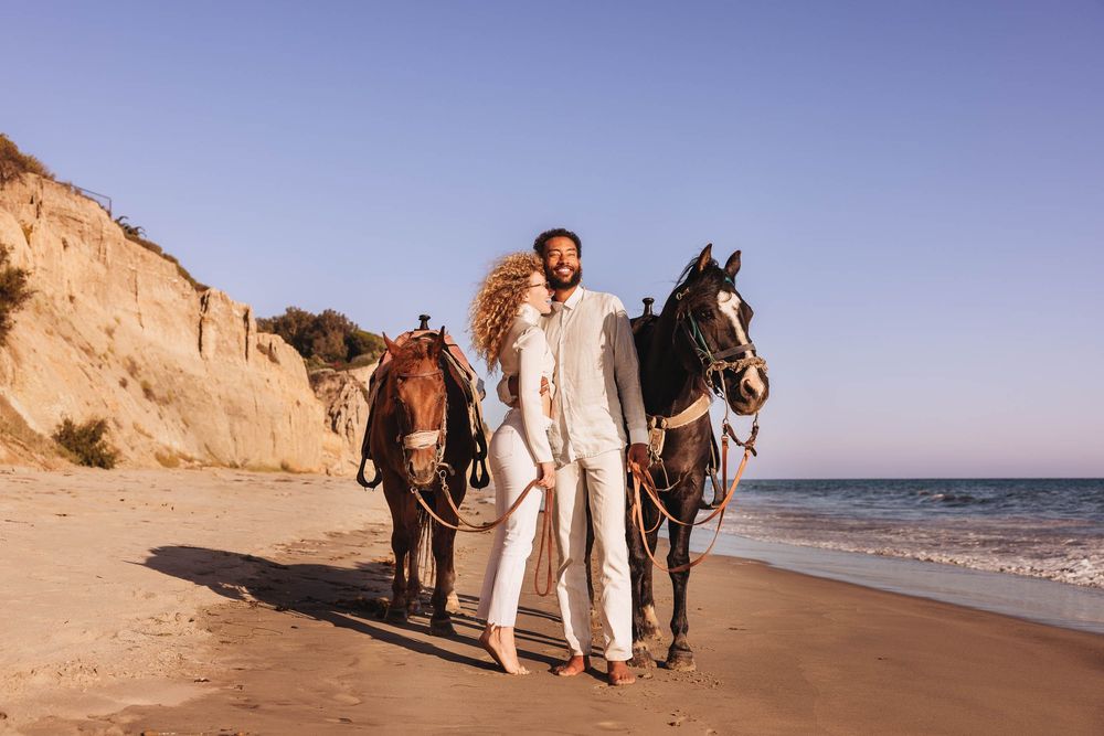 Couple in light colored outfits standing with two horses on a sandy beach with cliffs in the background.