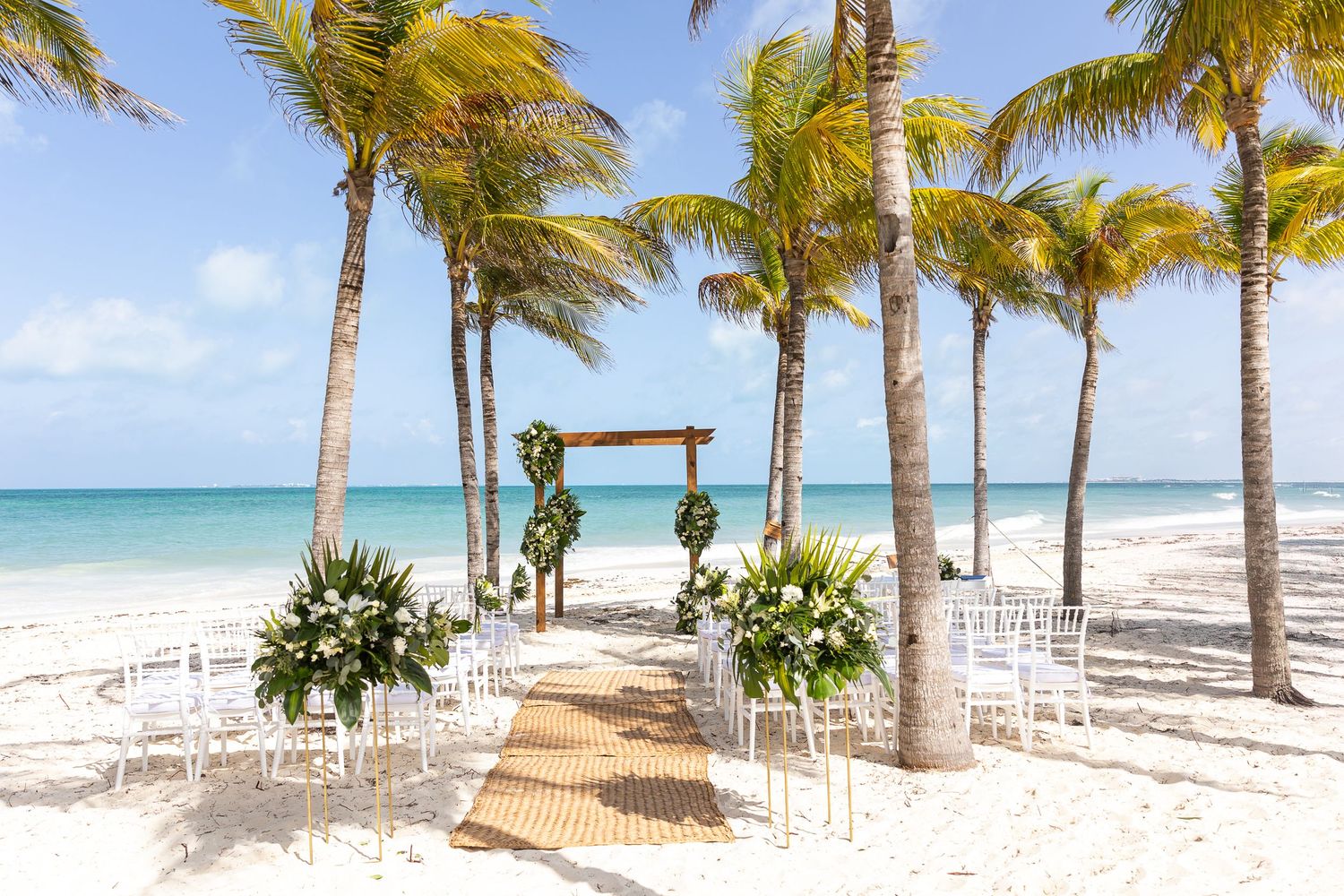 Tropical beach wedding setup with white chairs, wooden arch and palm trees along turquoise ocean.