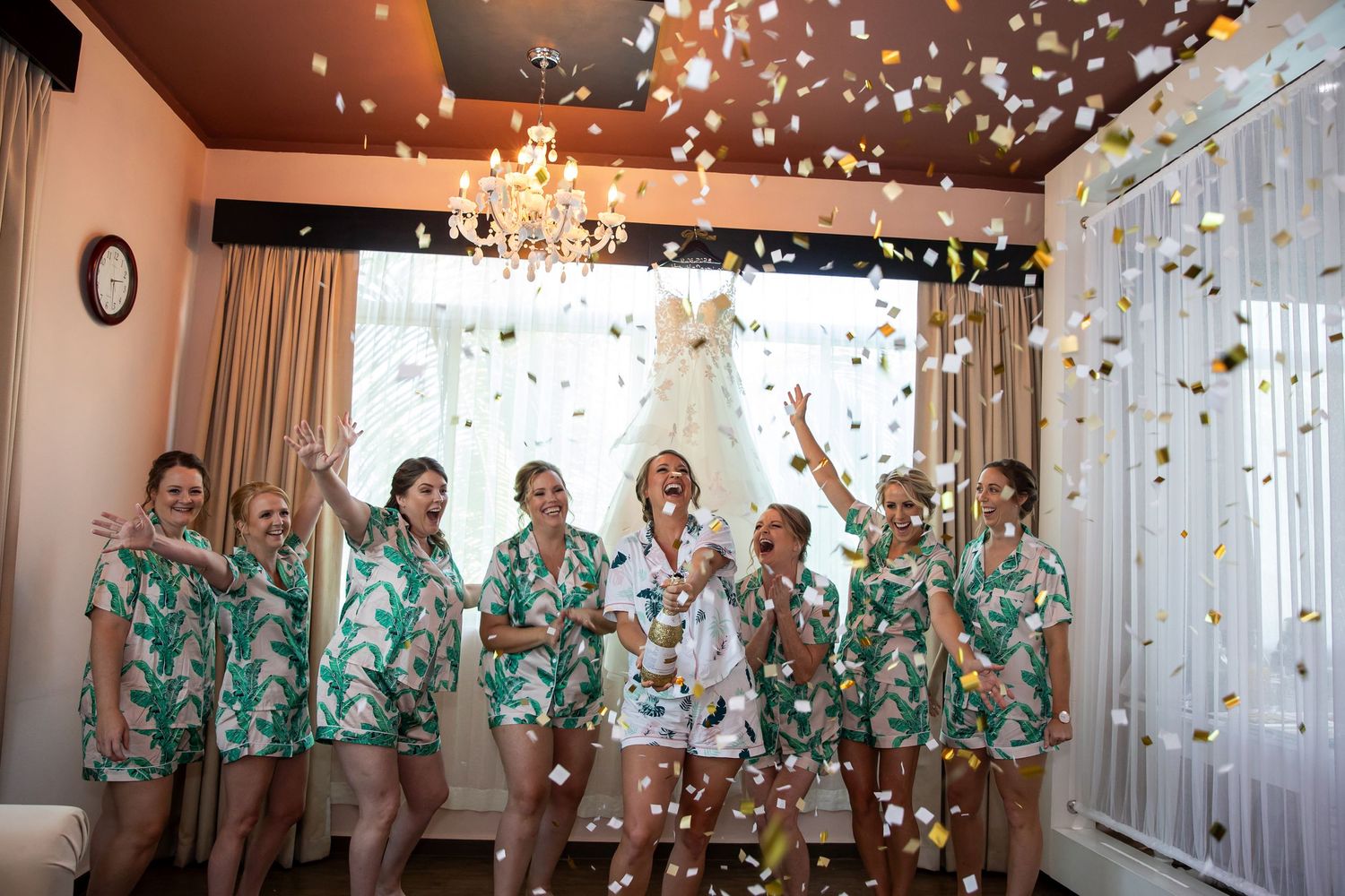 Bridesmaids in matching floral robes celebrate with confetti in an elegant getting-ready room.