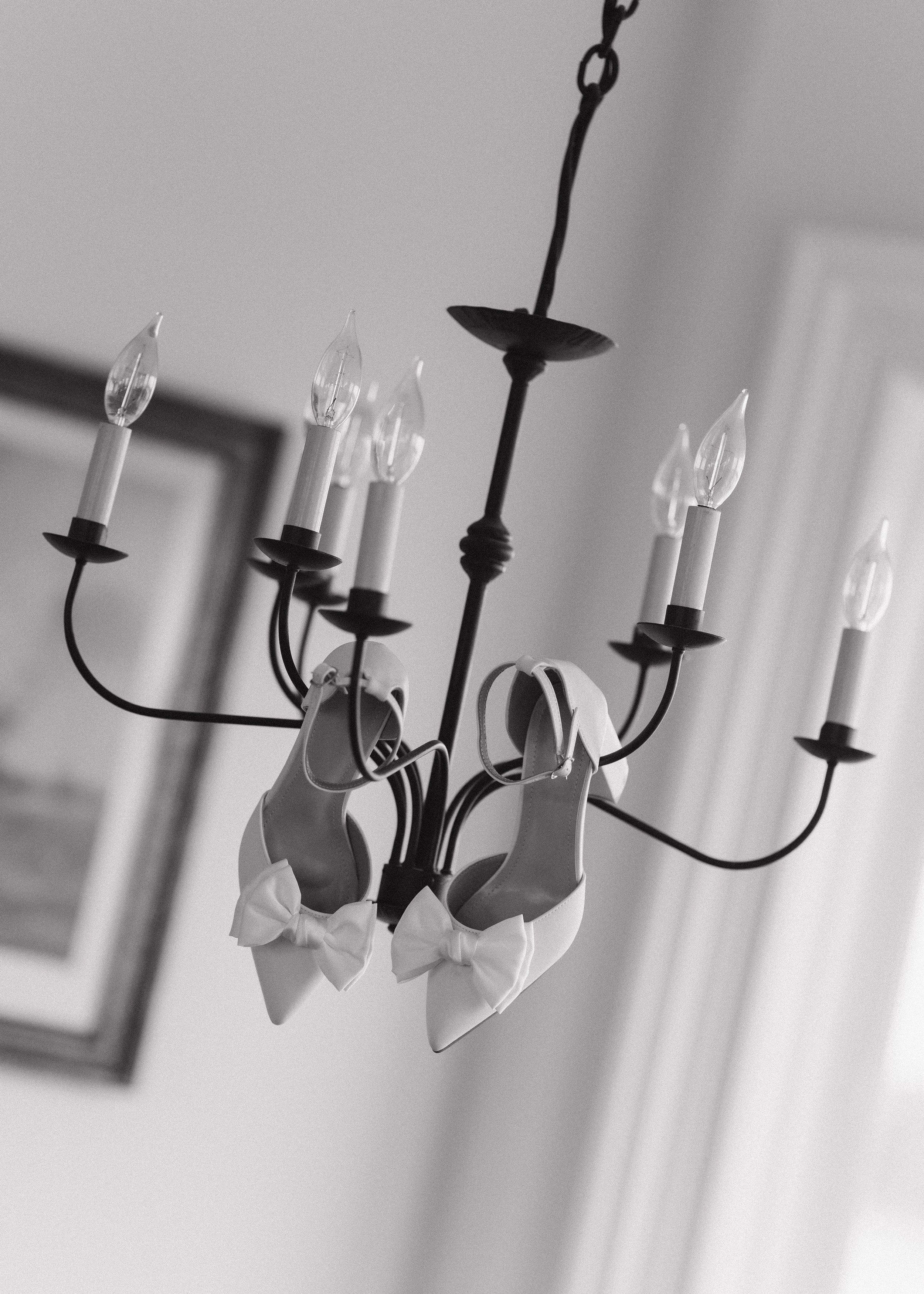 Black and white photo of white bridal heels with bows hanging from an elegant candelabra chandelier.