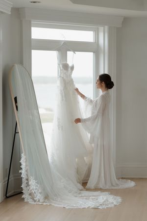 Bride in white robe admires lace wedding dress hanging in sunlit window with floor mirror beside her.
