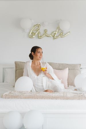 Smiling bride in white robe relaxes on bed holding orange juice surrounded by white balloons under gold bride sign.