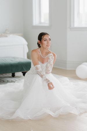 Bride in lace ballgown with sheer sleeves sitting gracefully on floor in bright white room.