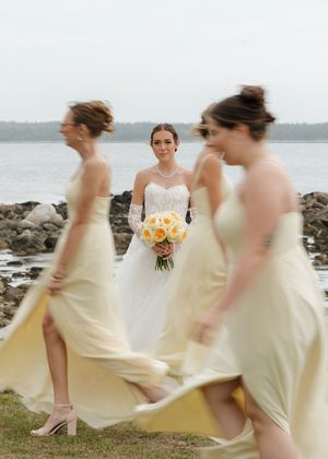Bride in white gown holds yellow bouquet as blurred bridesmaids in pale yellow dresses walk past a rocky shoreline.