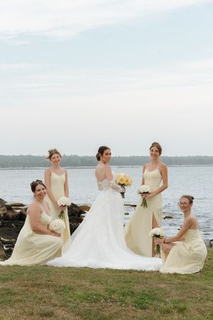 Bride with four bridesmaids in pale yellow gowns pose with bouquets by rocky waterfront on wedding day.