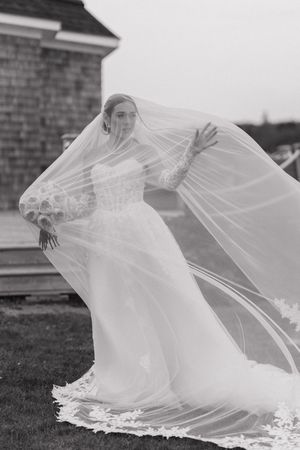 Black and white photo of bride in lace gown with flowing veil billowing in the wind outdoors.