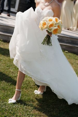 Bride in white strapless gown holding yellow rose bouquet, wearing white heels on green grass.