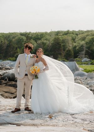 Bride and groom pose on rocky shore, her white ballgown veil flowing, holding yellow bouquet.