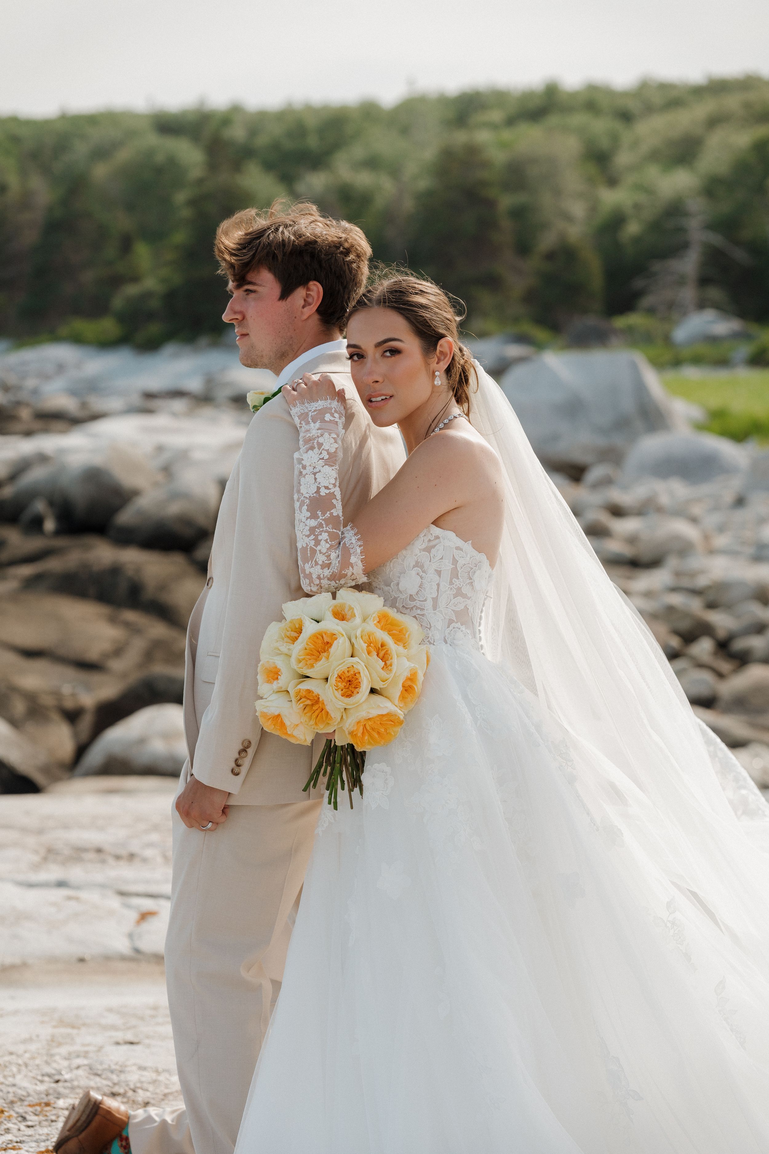 Bride in white ballgown with yellow bouquet leans on grooms shoulder near rocky coastal wedding location.