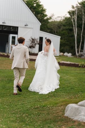 Bride in lace gown and groom in tan suit walking toward white barn venue at dusk.