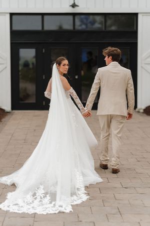Bride in lace wedding gown with cathedral veil holds hands with groom in tan suit walking toward venue doors.