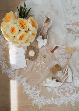 Elegant bridal flat lay with white heels, yellow roses, lace veil, wedding details card, and gold accessories on wood floor.
