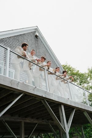 Groomsmen in white suits toast on a elevated deck outside a gray barn wedding venue.