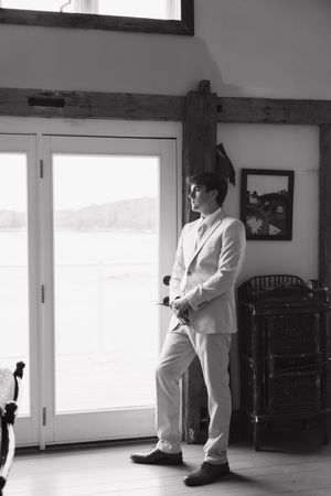 Black and white photo of a young man in a light suit standing by French doors in an elegant rustic interior.