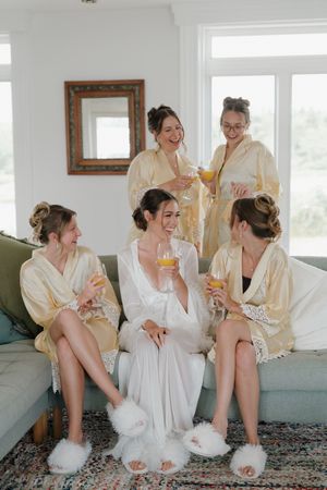 Bride in white robe with bridesmaids in gold robes toasting with mimosas on a couch before the wedding.