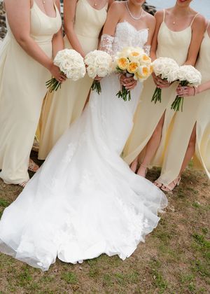 Bride in white lace gown with bridesmaids in pale yellow dresses holding white and cream rose bouquets outdoors.