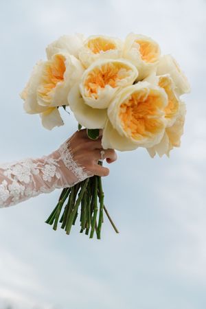 Bride holding a bouquet of yellow and peach garden roses against a blue sky.