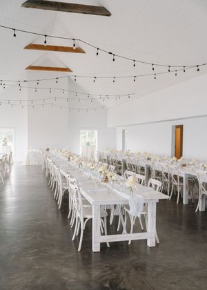 Elegant white wedding reception hall with long farmhouse tables, white chairs, string lights, and soft floral centerpieces.