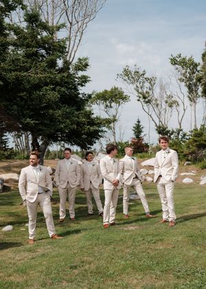 Groom and groomsmen in matching cream suits walking outdoors on green grass with trees and blue sky.