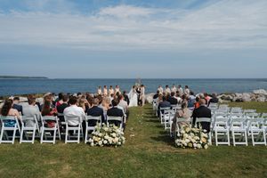 Outdoor wedding ceremony by the ocean with guests seated in white chairs on a green lawn.
