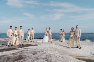 Wedding party poses on rocky shoreline in cream and beige attire with ocean backdrop on a clear day.