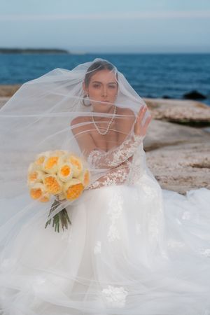 Bride in lace wedding gown and flowing veil holds yellow rose bouquet by the ocean.