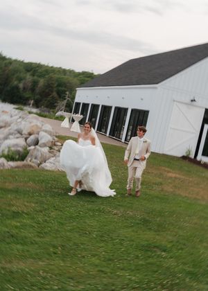 Bride and groom walking joyfully across green lawn beside a white barn wedding venue.