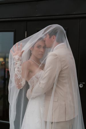 Bride and groom share intimate moment beneath flowing white veil, groom in tan suit kissing brides cheek.