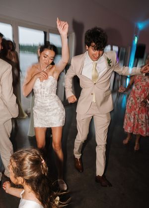 Bride in white mini dress and groom in beige suit dancing joyfully at their wedding reception.