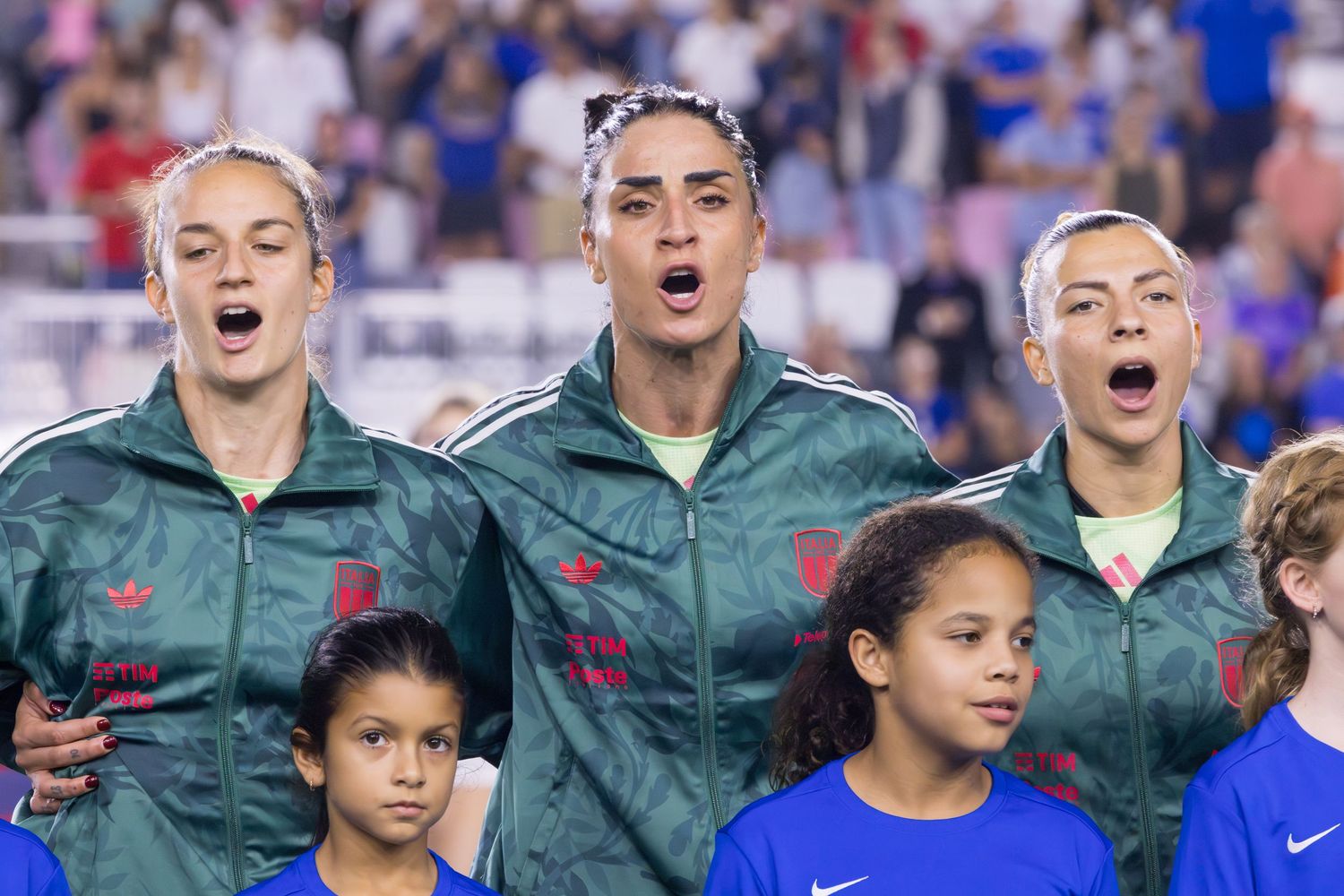 Three female soccer players in green jackets sing the national anthem at an international match.