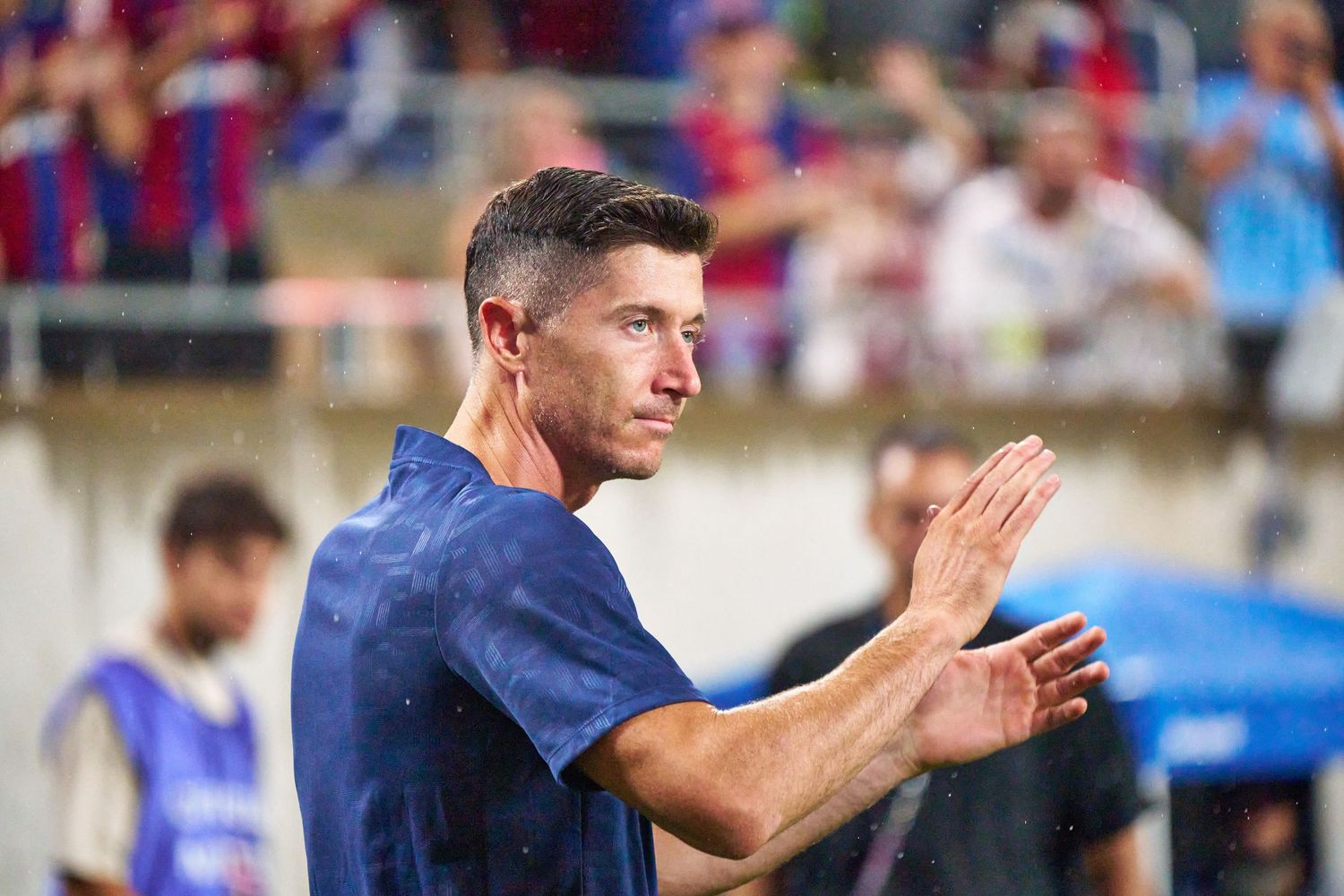 A person in a navy blue shirt applauds while standing on a sports field with spectators in the background.