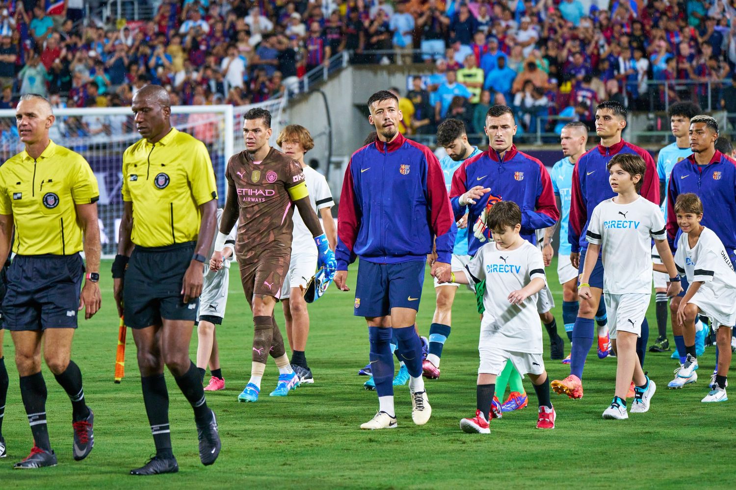 Soccer players and match officials walk onto the field with young player escorts before a match at a packed stadium.