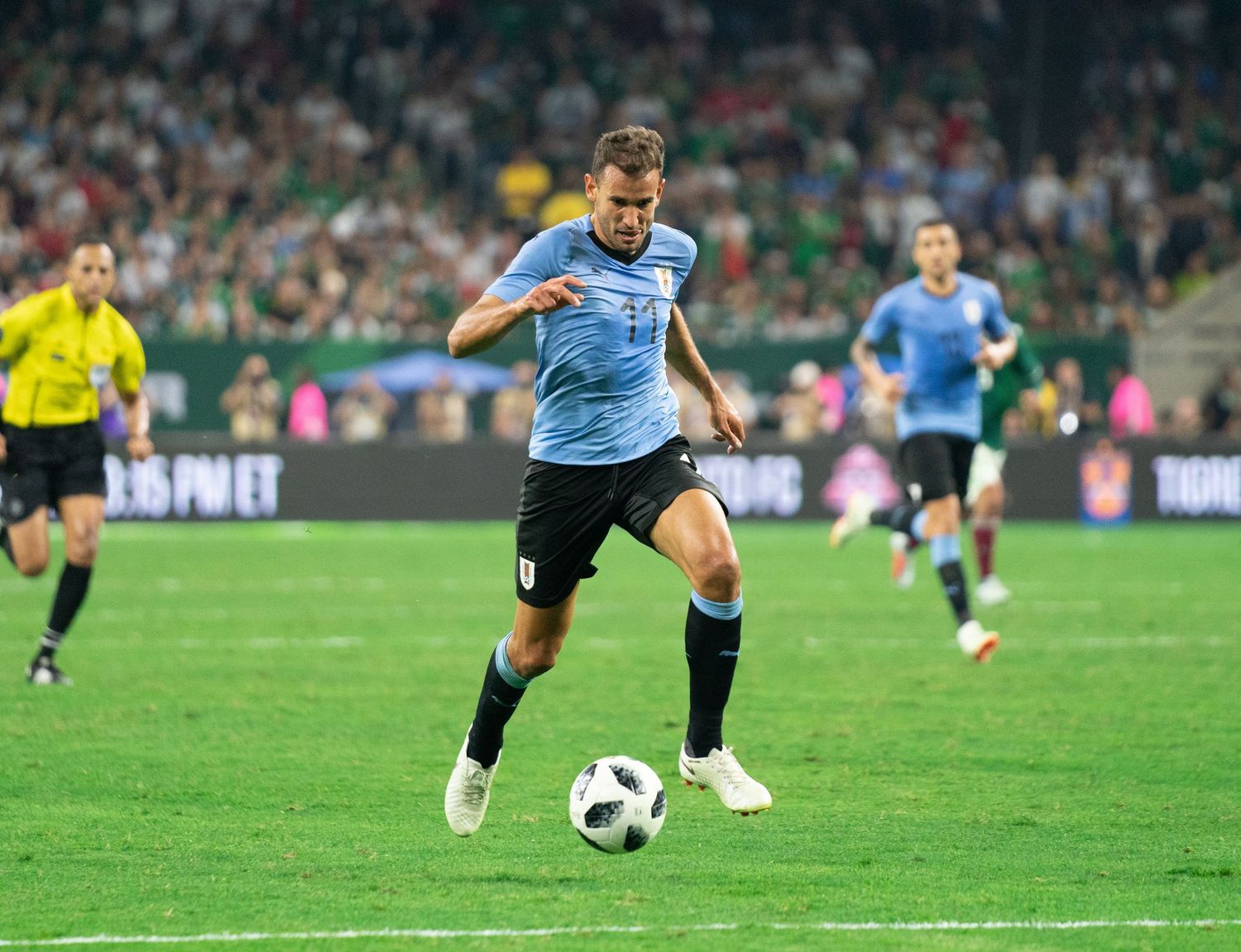 Soccer player in light blue jersey dribbles ball on grass field during competitive match.