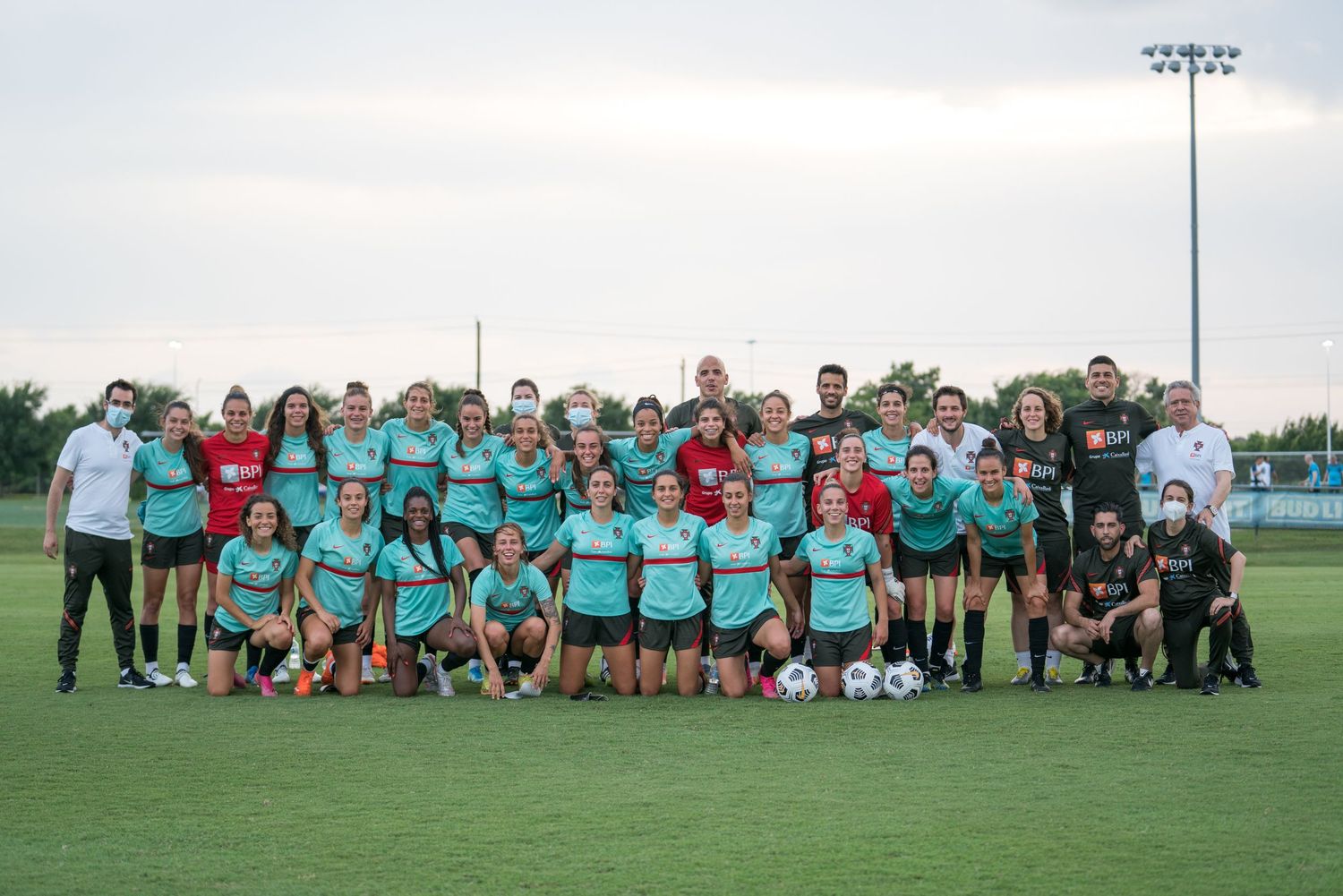 Large group of soccer players in turquoise jerseys pose together on a sunny field for a team photo.