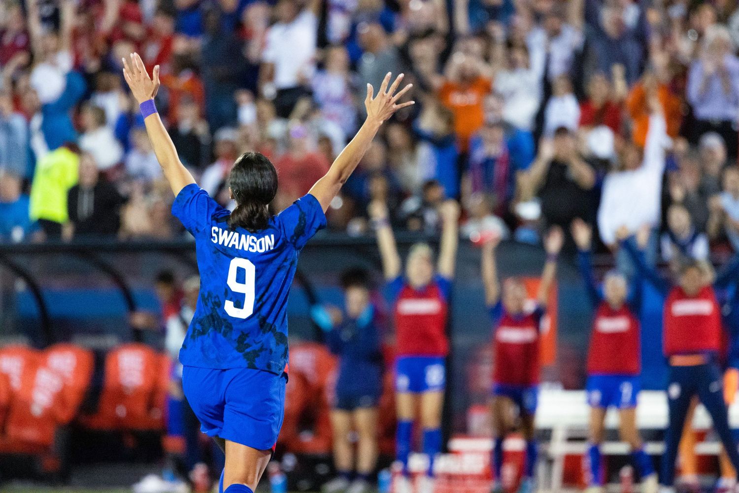 Soccer player Swanson #9 in blue uniform raises arms celebrating on field during a match.