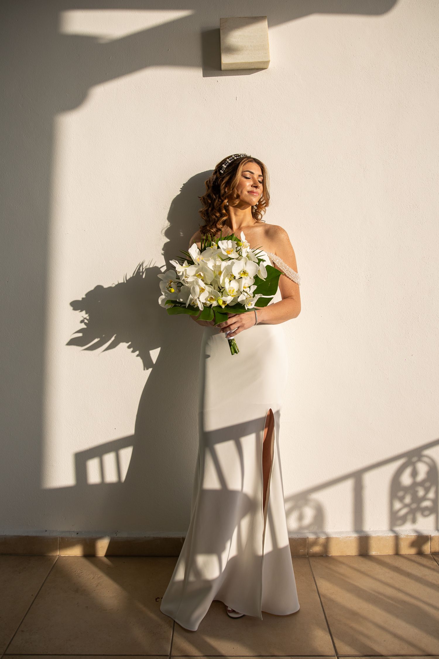 Elegant bride in white gown holding white floral bouquet casting dramatic shadow against white wall.
