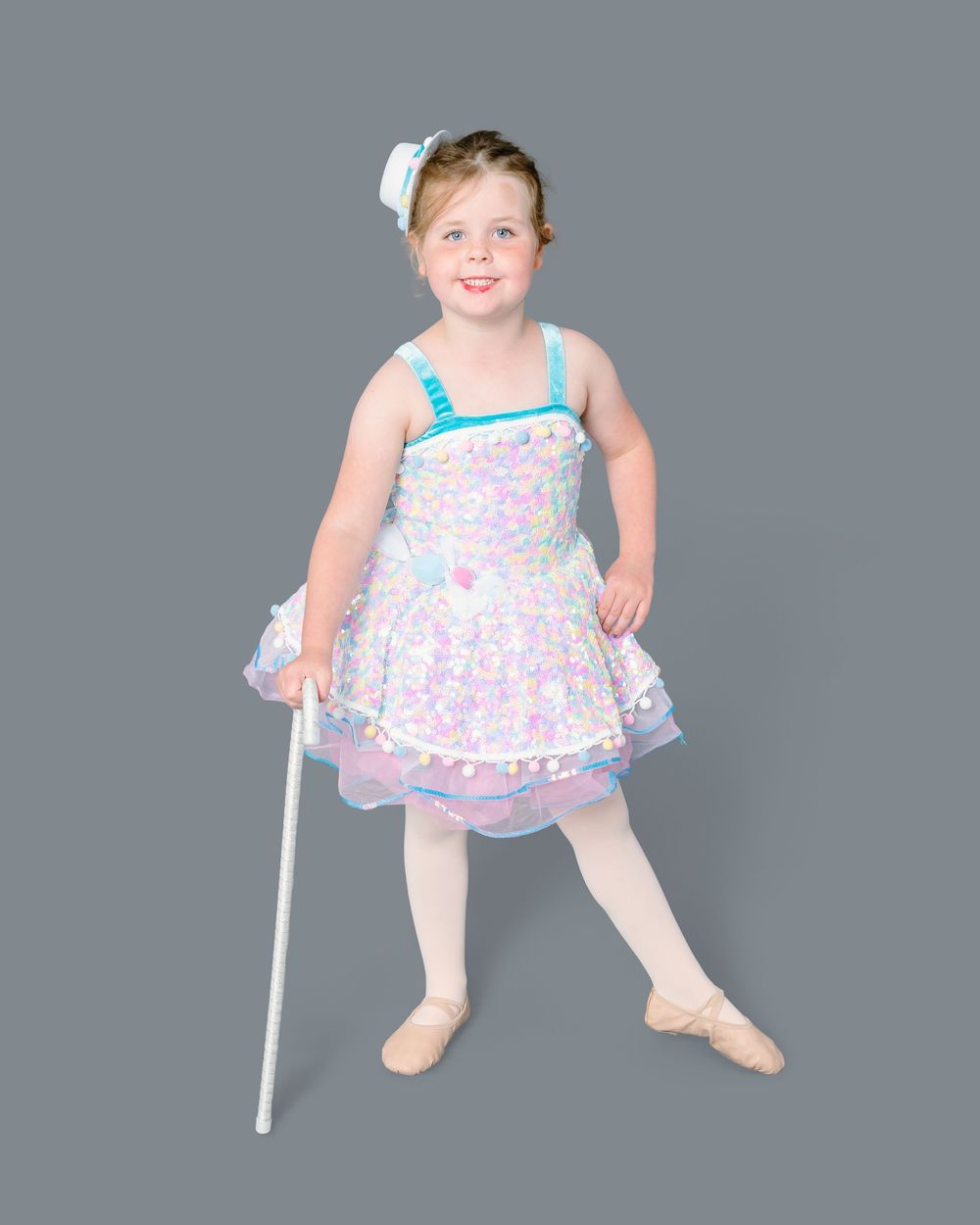 A dancer in a pink and blue tutu dress with white tights performs a graceful pose against a gray backdrop.