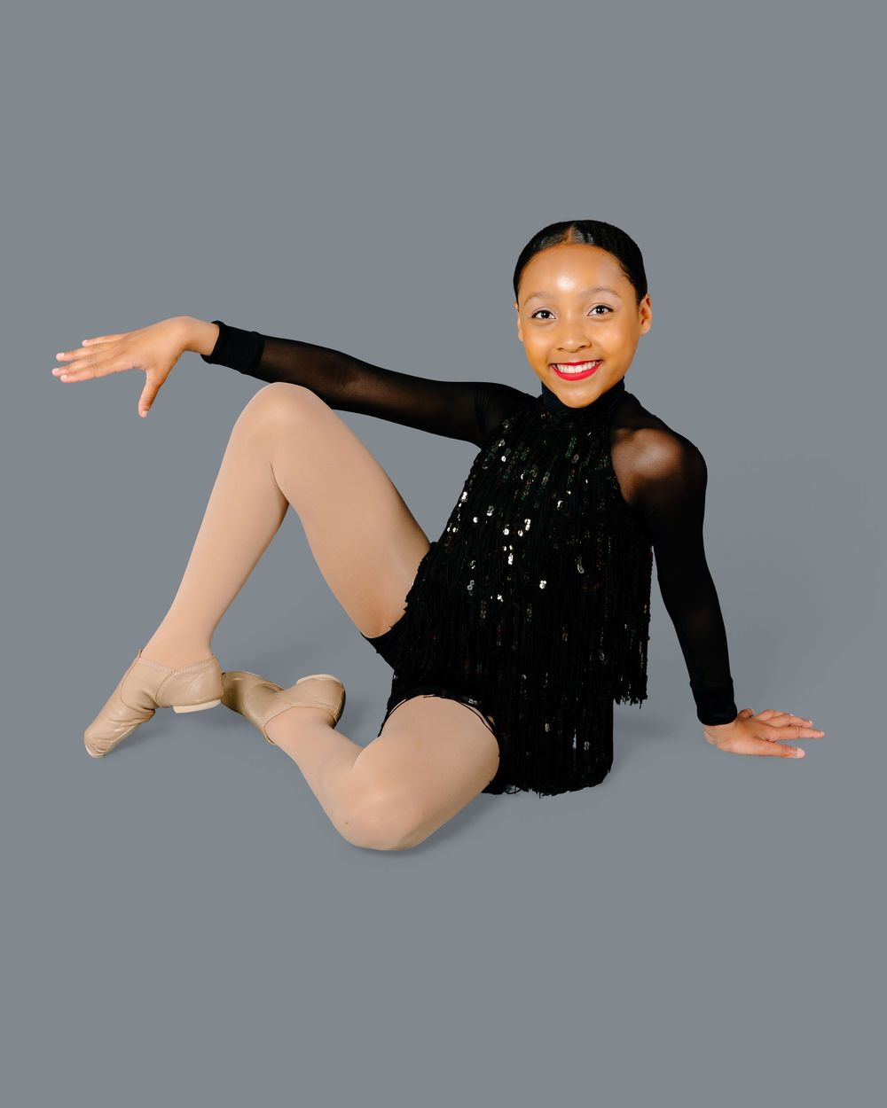 Dancer in black sequined leotard strikes a seated pose with pointed leg against gray backdrop.