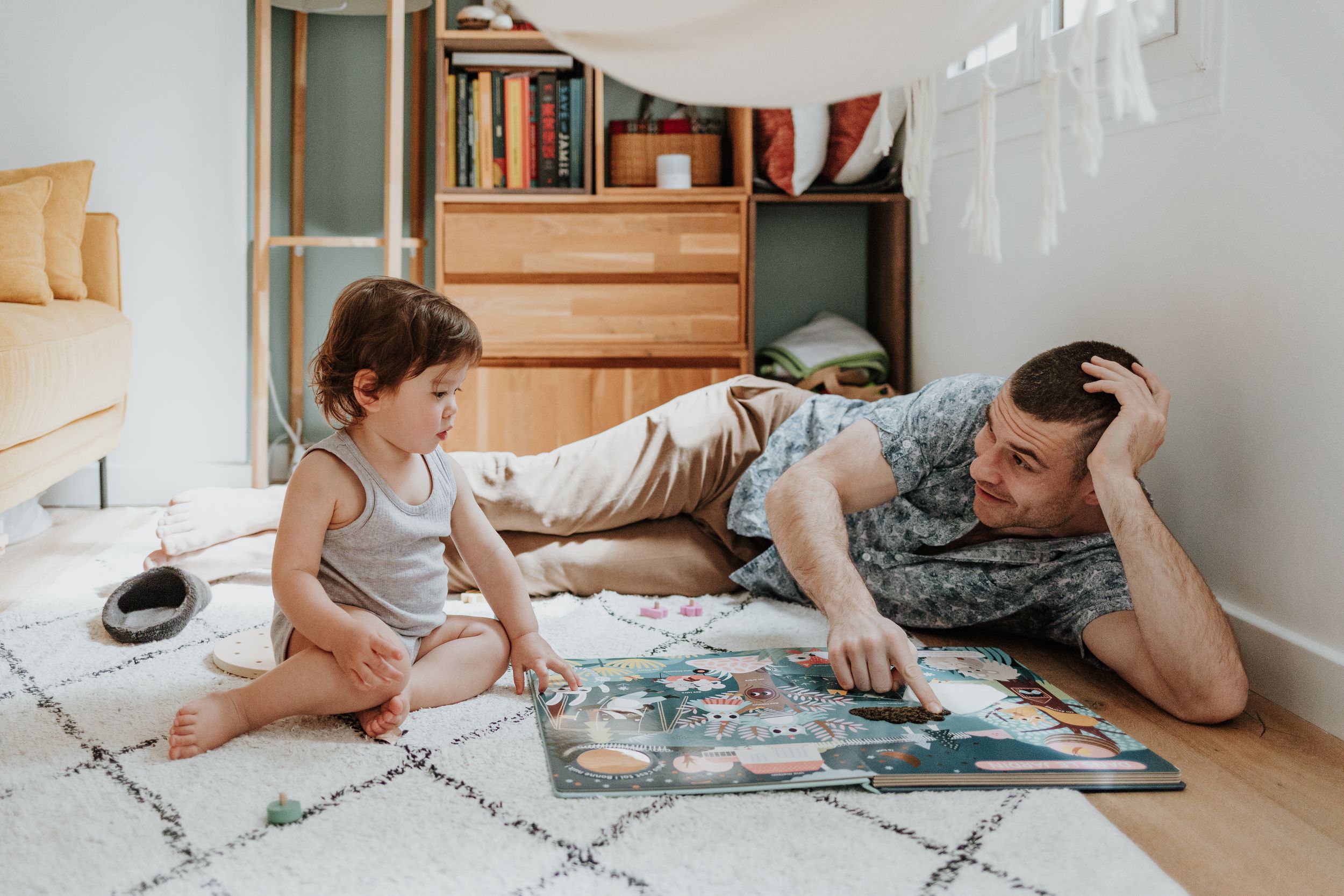 Two people lay on a cozy carpet reading a book together in a room with built-in wooden bookshelves.