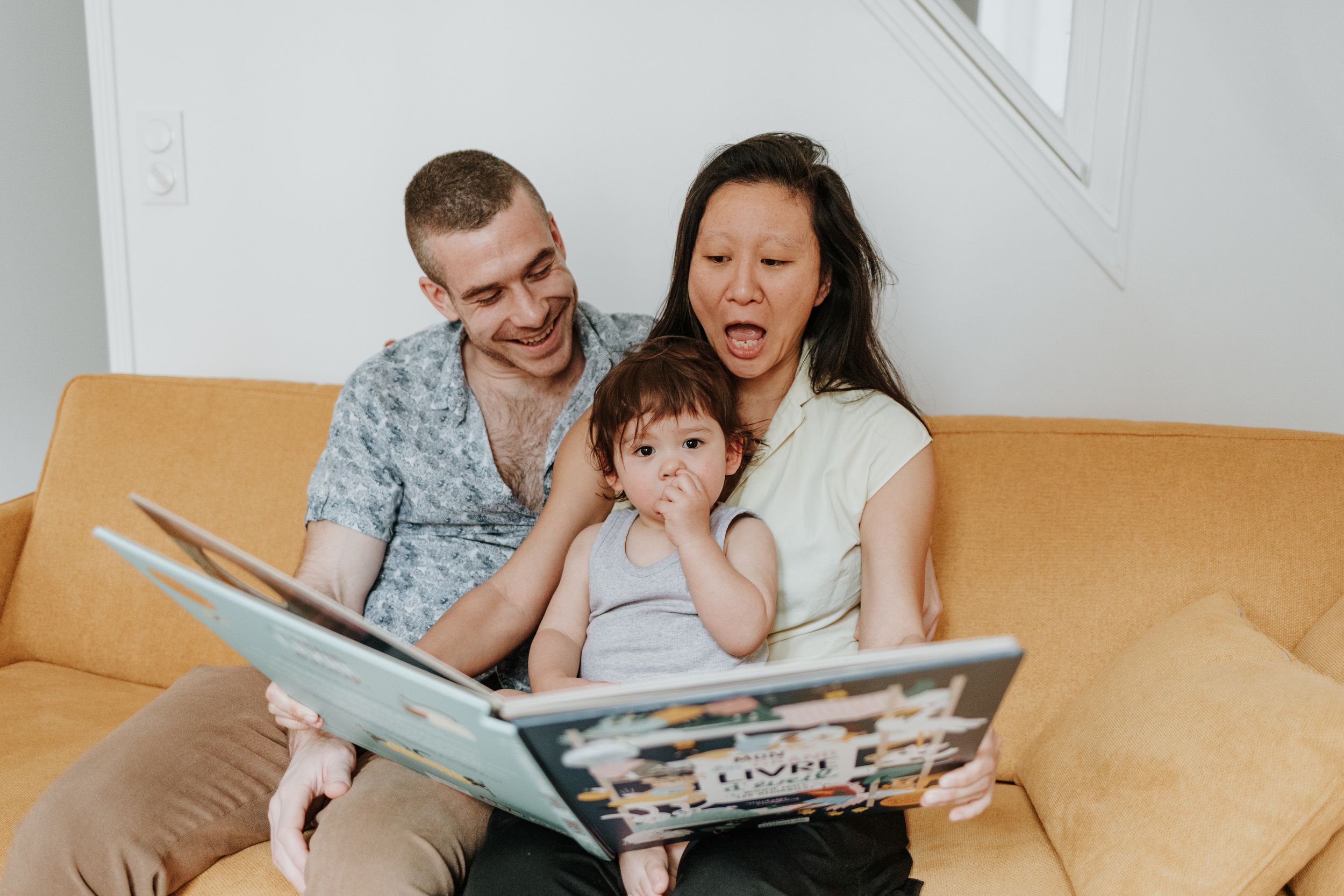 A family sits together on a yellow couch reading a large picture book during storytime at home.