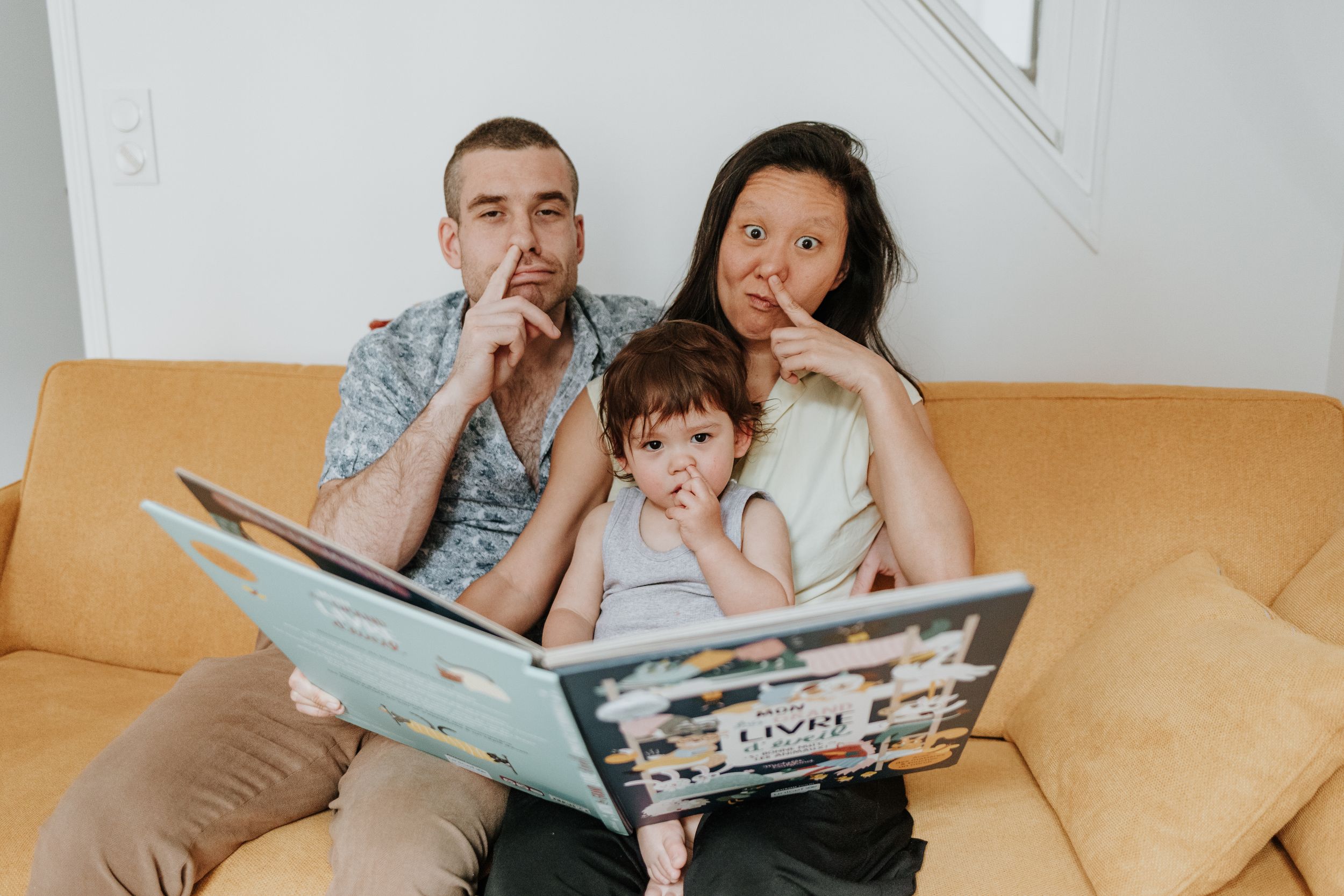 Family sitting close together on orange couch reading a picture book and making silly faces.