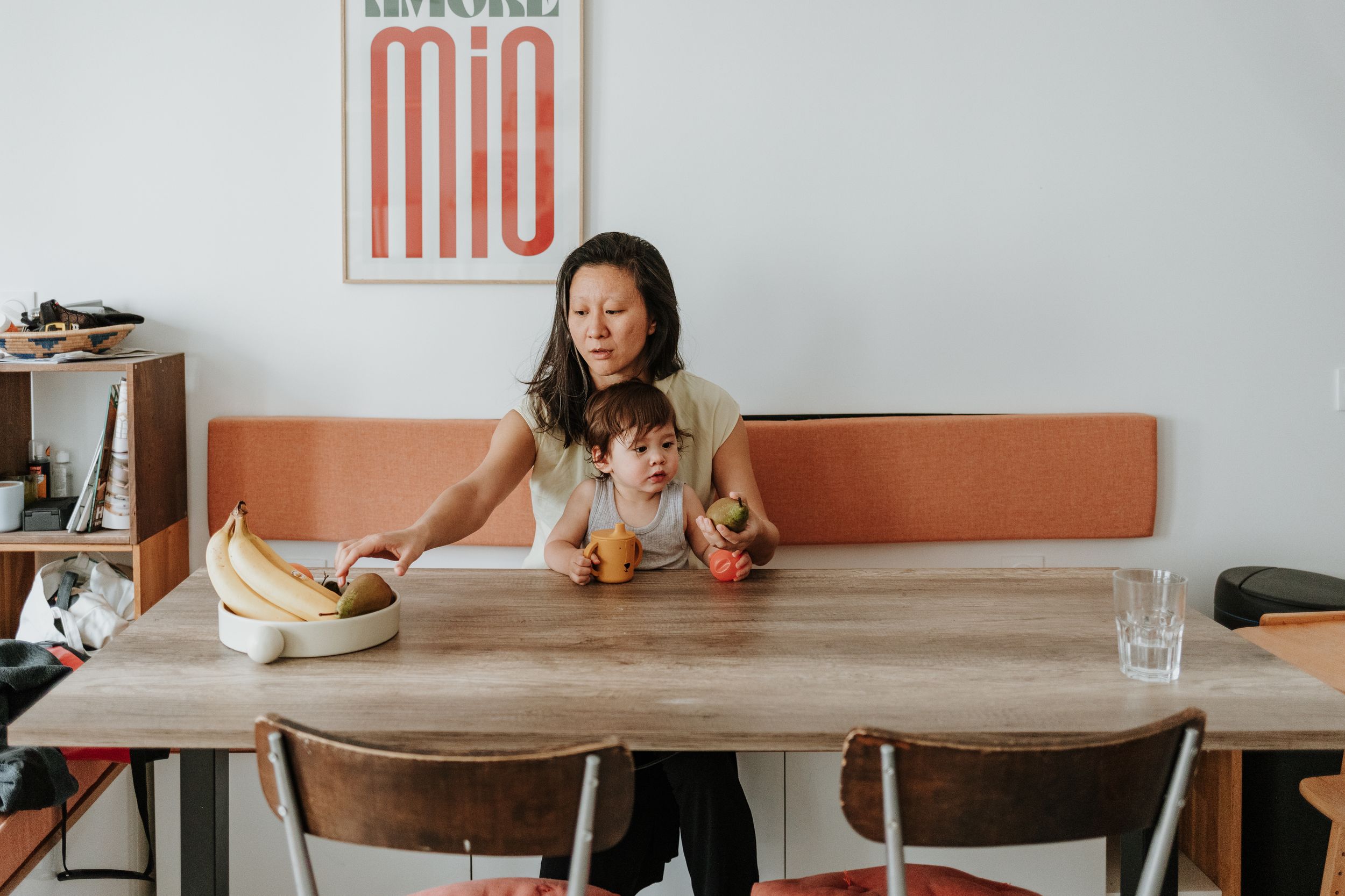 A person works at a wooden table with books and drinks, sitting beneath vintage wall art in a minimalist space.