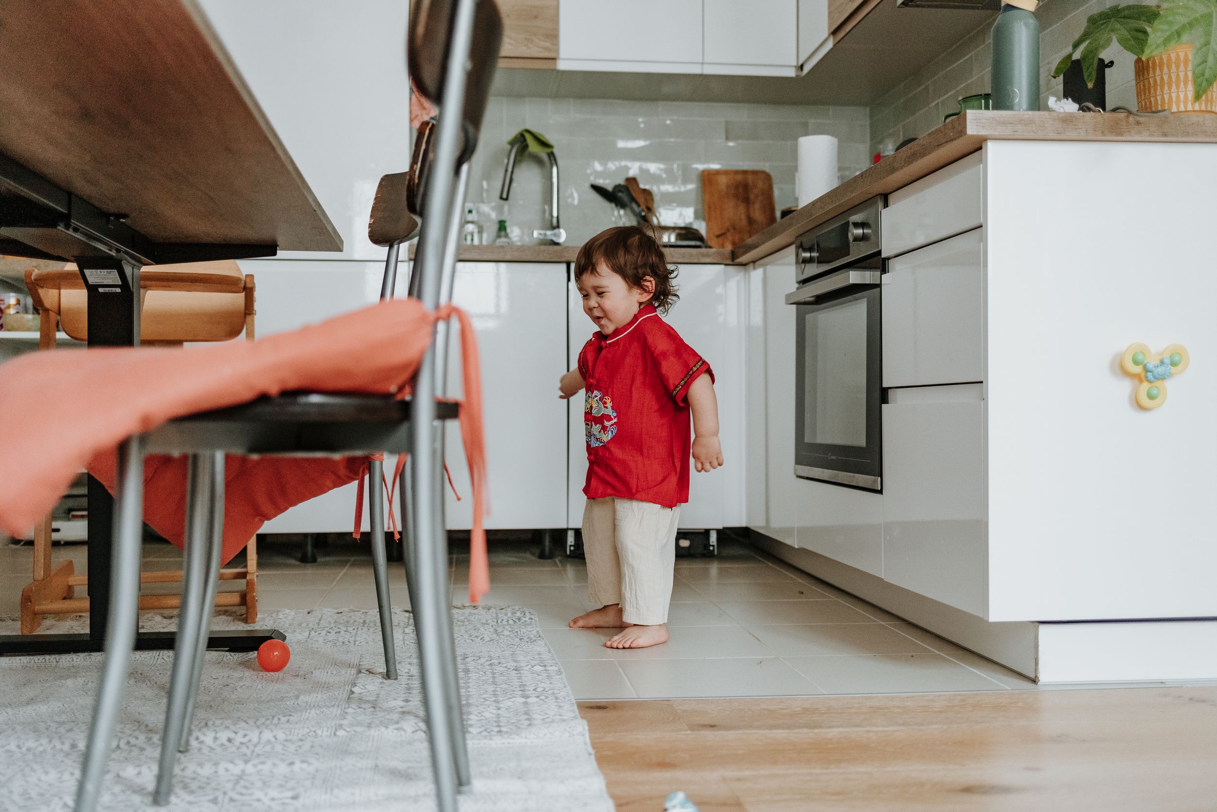 A small child in a bright red sweater stands at attention in a modern kitchen with hardwood floors.