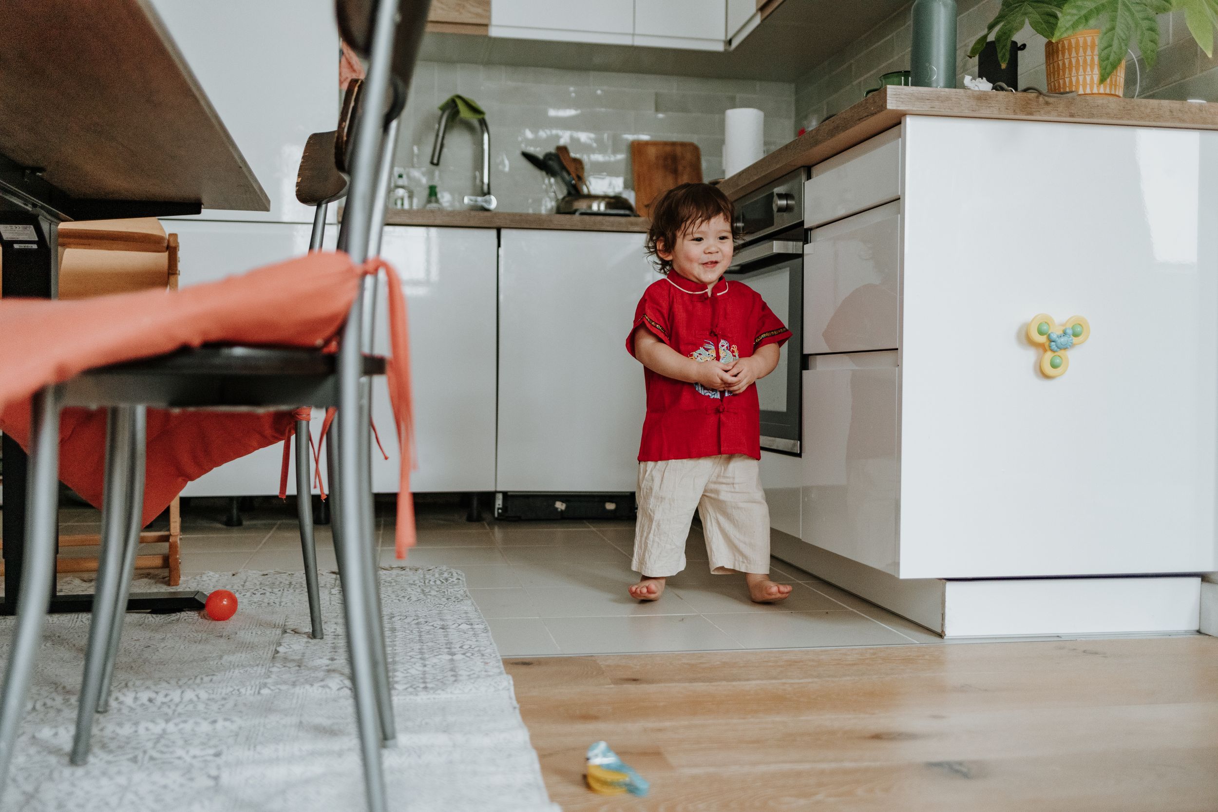 Small child wearing red shirt and beige shorts stands in modern kitchen with hardwood floors and white cabinets.