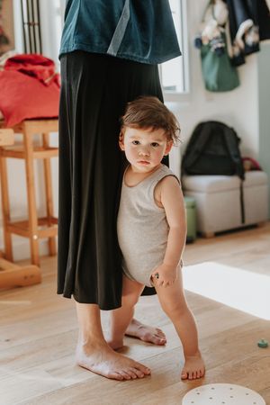 A toddler wearing a beige tank top bodysuit stands barefoot next to an adult in a long black skirt on hardwood flooring.