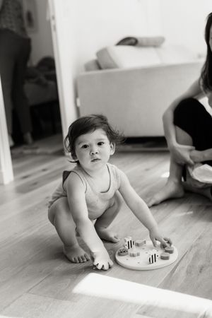 A small child in a white sleeveless top sits on wooden flooring playing with a colorful toy in natural lighting.